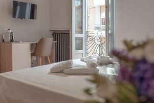 Habitación de hotel con cama, escritorio, silla, televisor y balcón con barandilla.