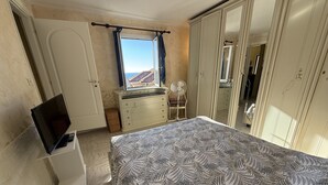 A bedroom with a bed, a TV, a dresser, a mirror, and a window with a view of buildings. - The Piano Apartment (Imperia)