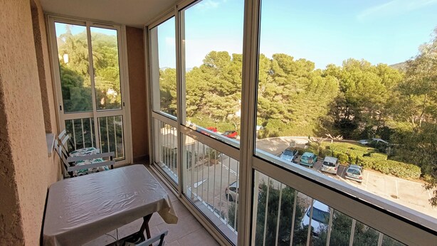 A balcony with a table and chairs, large windows offering a view of a parking area and trees. - Apartment with balcony near beach (Six-Fours-les-Plages)