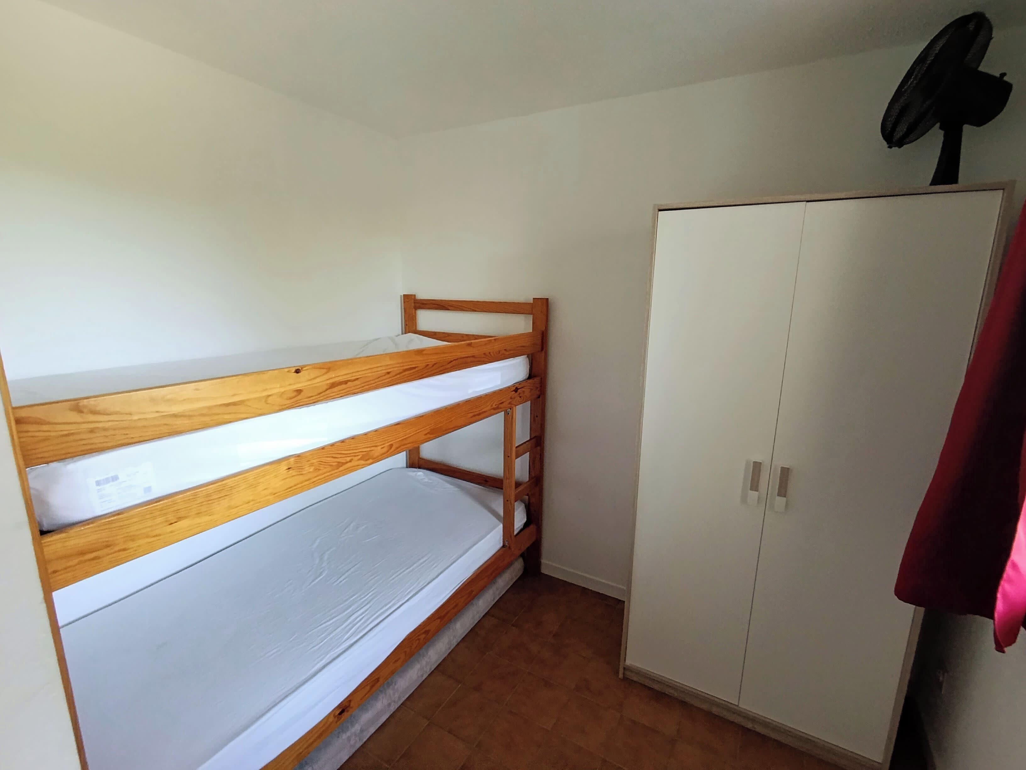 Una cama litera con estructura de madera y colchón blanco, un armario de dos puertas y un ventilador de techo.