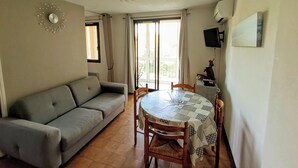 A living room with a grey sofa, a round wooden dining table with chairs, a ceiling fan, a wall clock, and a TV mounted on the wall. - Apartment with balcony near beach (Six-Fours-les-Plages)