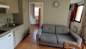 A compact living space with a kitchenette, a grey sofa, and a wooden chair. A decorative clock is mounted on the wall. - Apartment with balcony near beach (Six-Fours-les-Plages)