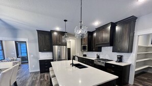 Une cuisine moderne avec des meubles en bois foncé, un grand îlot central doté d’un évier, des appareils électroménagers en acier inoxydable et des luminaires suspendus.