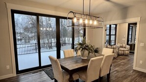 Un coin repas avec une table et des chaises en bois, un luminaire suspendu et une vue sur un espace extérieur enneigé, visible à travers de grandes portes-fenêtres coulissantes.