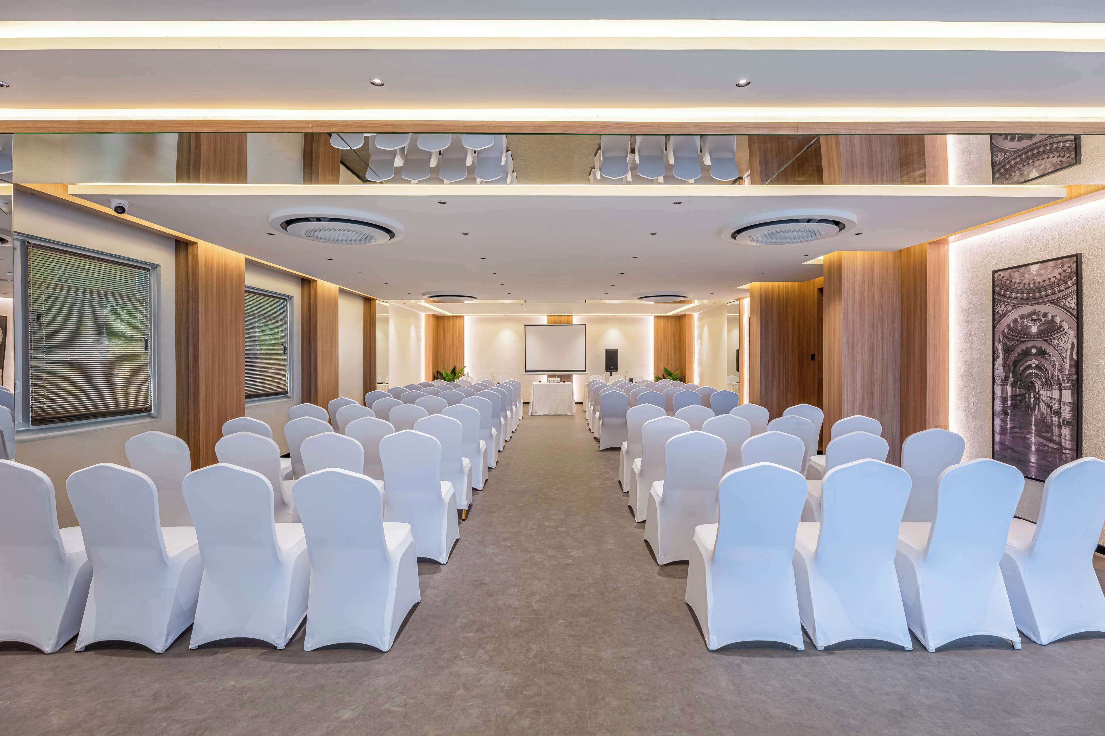 Un salón de conferencias con filas de sillas blancas, un escenario al frente y una pantalla grande.