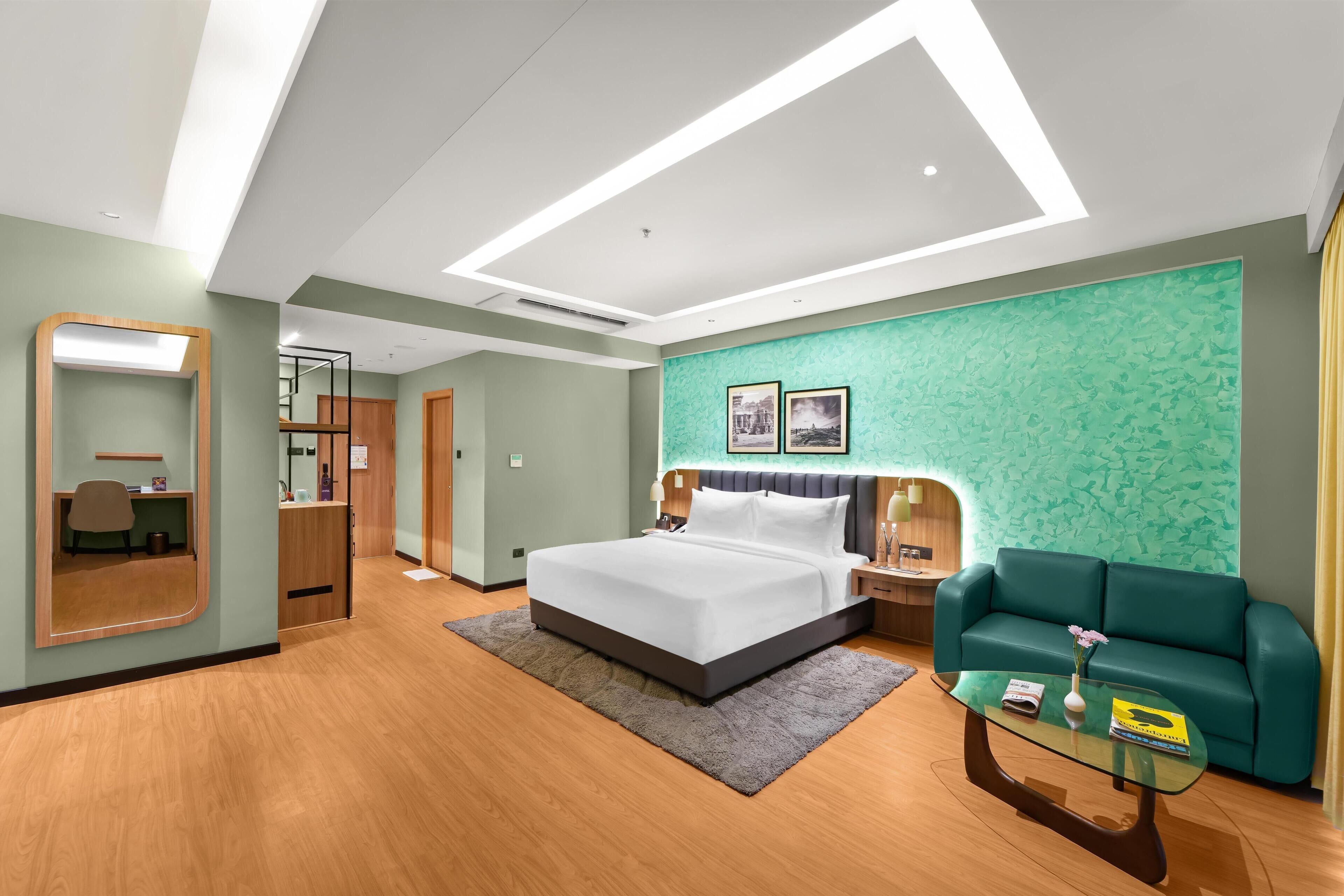 A modern hotel room with a large bed, a seating area with a sofa and coffee table, a desk with a chair, and a mirror.