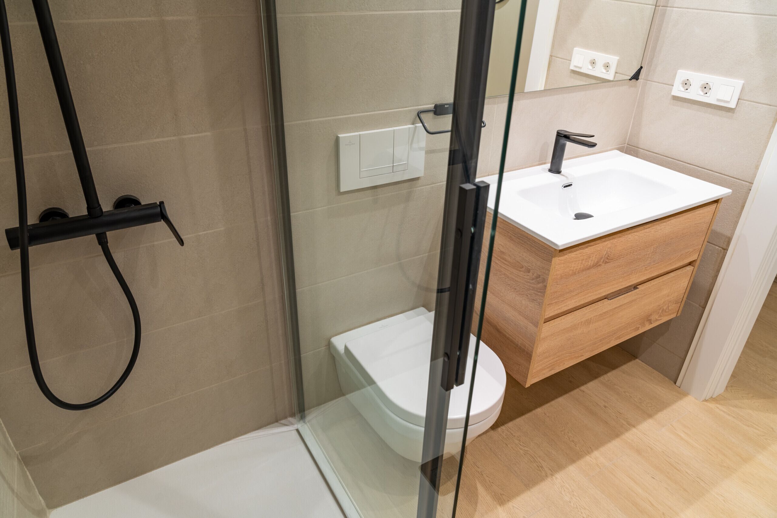Un baño moderno con cabina de ducha de vidrio, un inodoro blanco, un mueble de tocador de madera con lavamanos y un espejo sobre el mueble.
