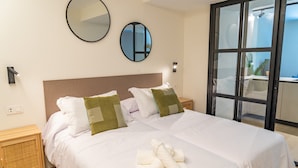 Uma cama de casal bem arrumada com roupa de cama branca e almofadas verdes, um espelho redondo e uma janela com vista para o exterior.