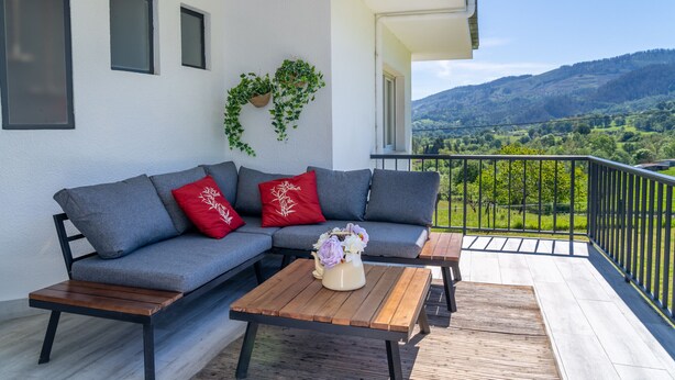 Een balkon met een grijze bank, rode en witte kussens, een houten tafel met een bloevas en uitzicht op groene heuvels.