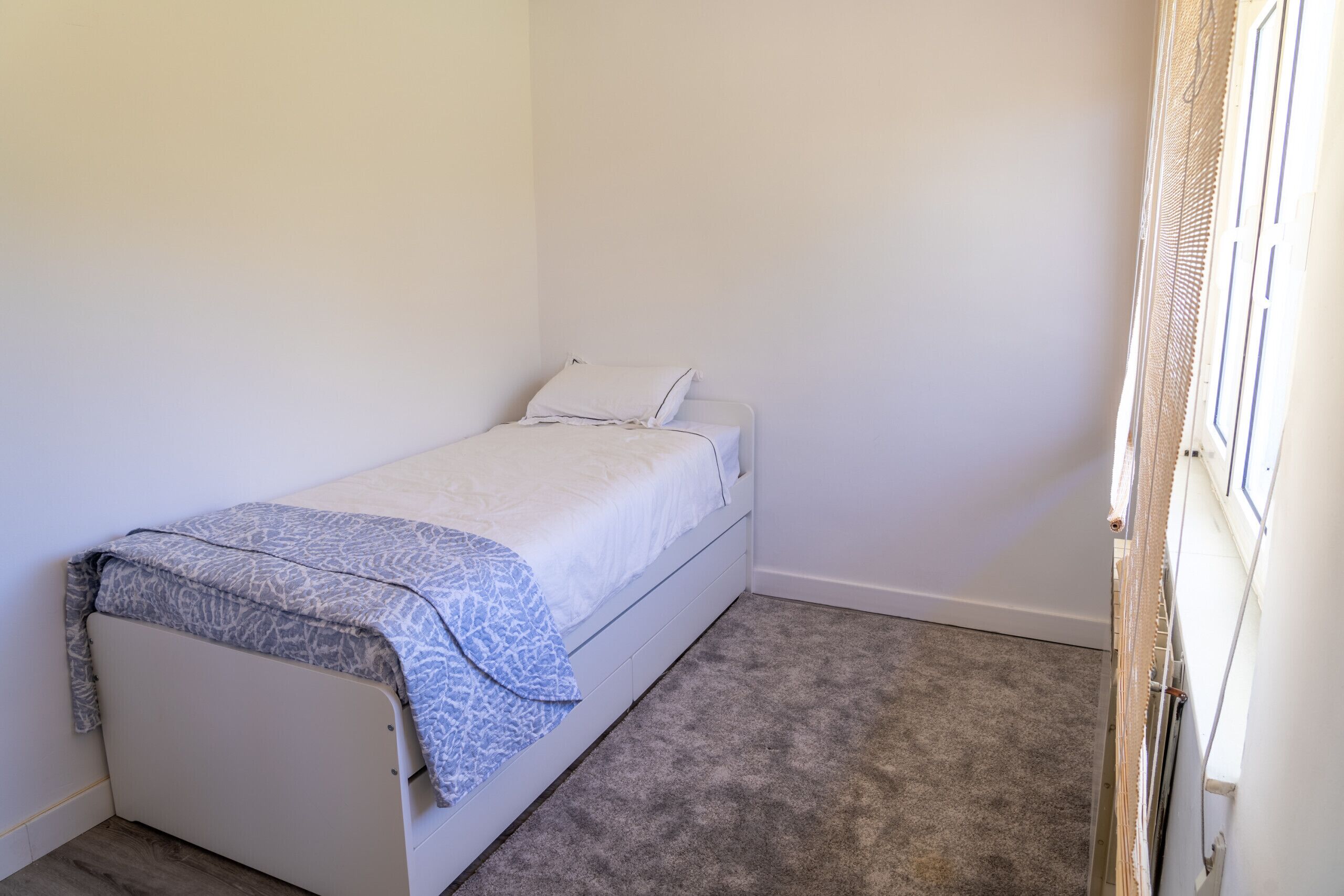 Een kleine kamer met een enkel bed, een grijs vloerkleed, witte wanden en een raam met een witte zonwering.