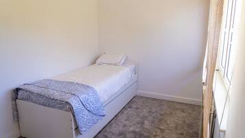 Een kleine kamer met een enkel bed, een grijs vloerkleed, witte wanden en een raam met een witte zonwering.