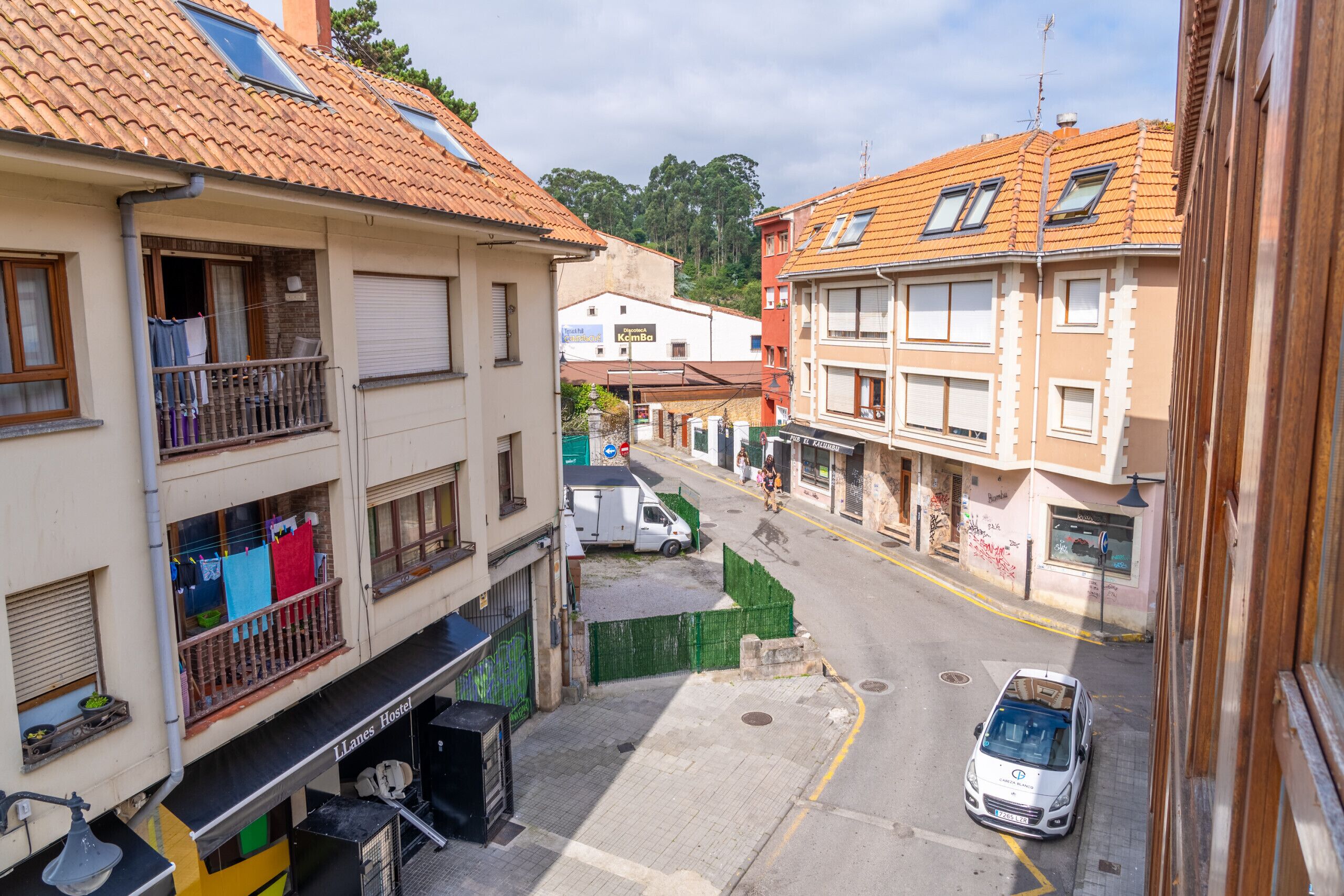 Una calle estrecha flanqueada por edificios, con una camioneta blanca estacionada en la acera y un coche aparcado en la calle.