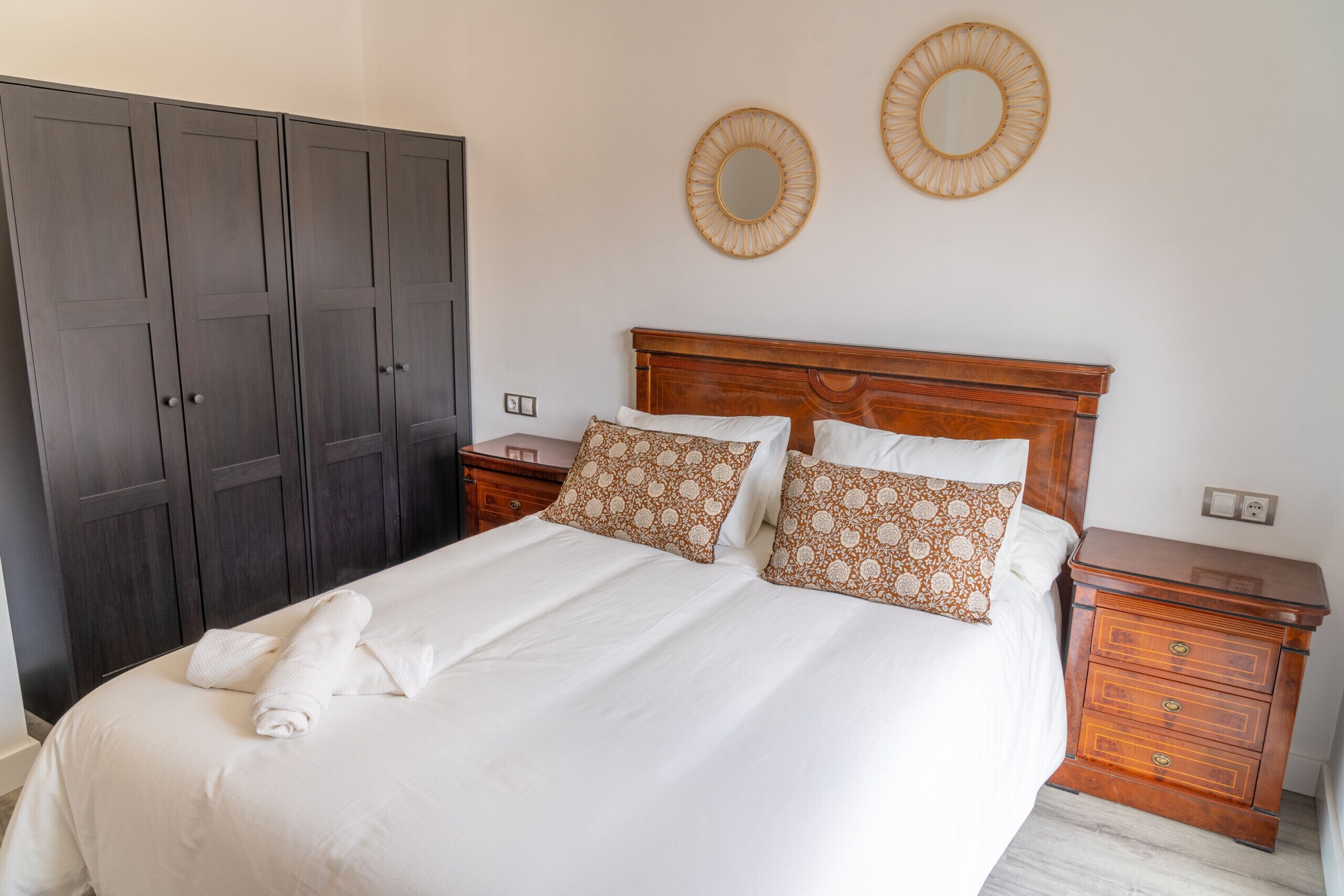 Una cama bien tendida con sábanas blancas y almohadones estampados, una mesita de noche de madera con cajones, y un armario oscuro.