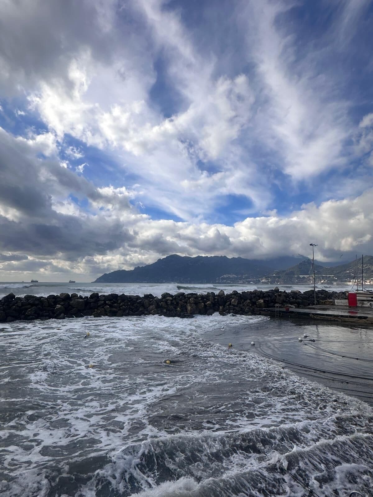 Paisaje costero con olas rompiendo sobre una costa rocosa, cielo despejado con algunas nubes y una cadena montañosa distante.