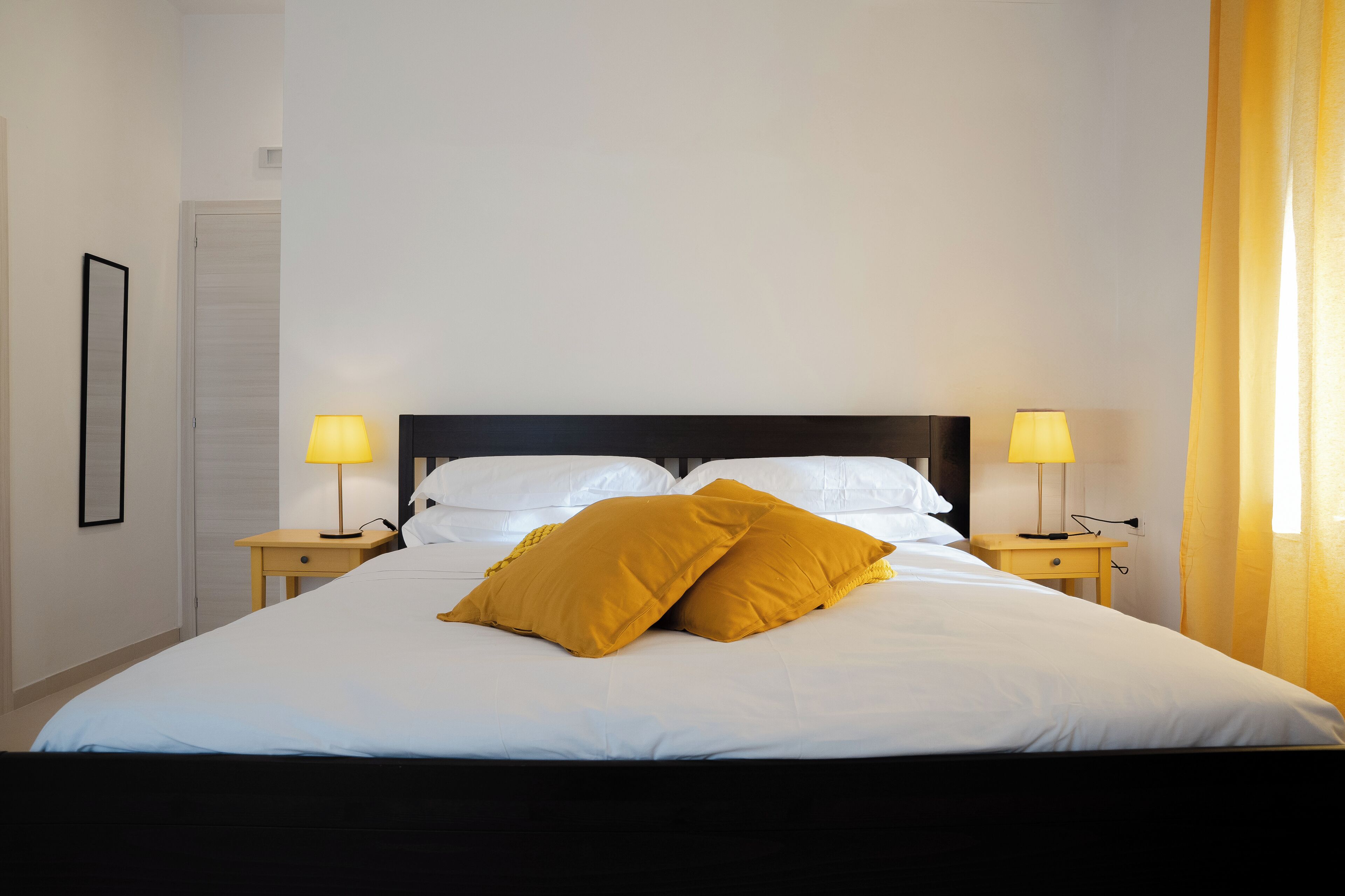Una cama bien hecha con sábanas blancas y dos almohadas amarillo mostaza, flanqueada por mesitas de noche con lámparas, en una habitación con una puerta blanca y paredes lisas.