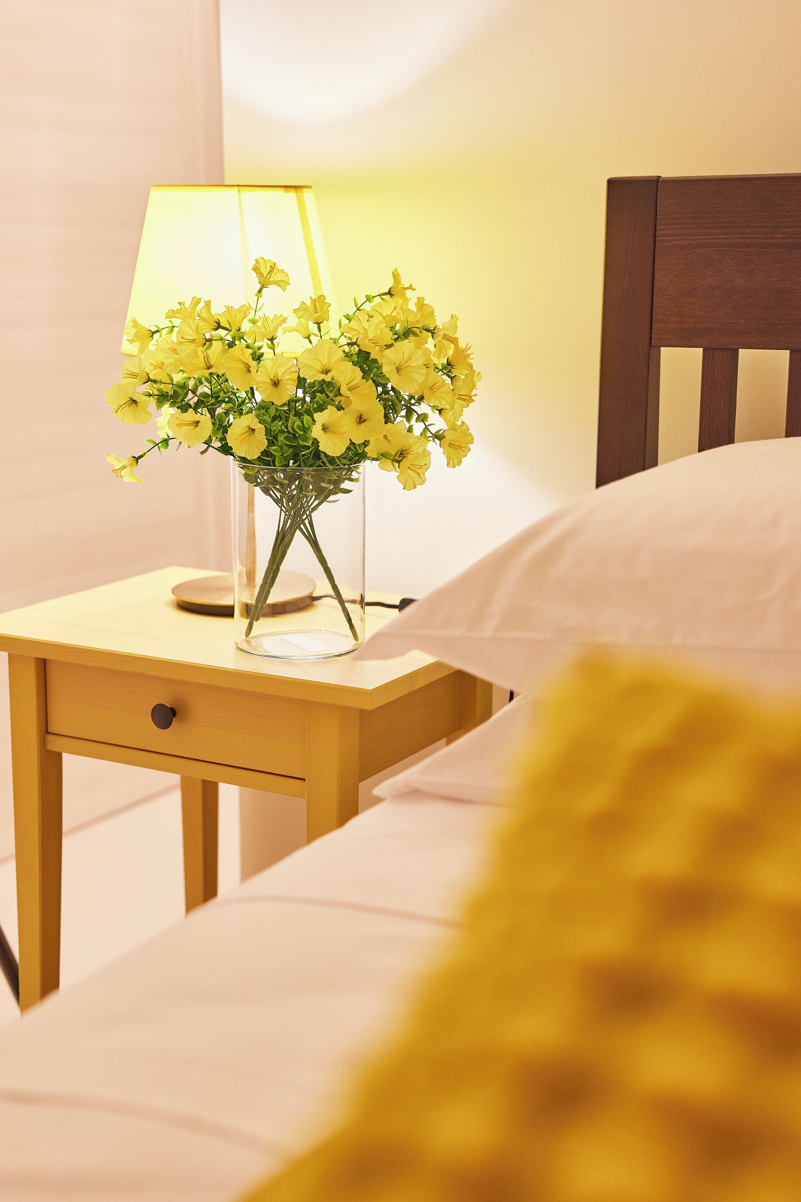 Una cama con cabecera de madera, una mesita de noche con una lámpara, y un florero transparente con flores amarillas.