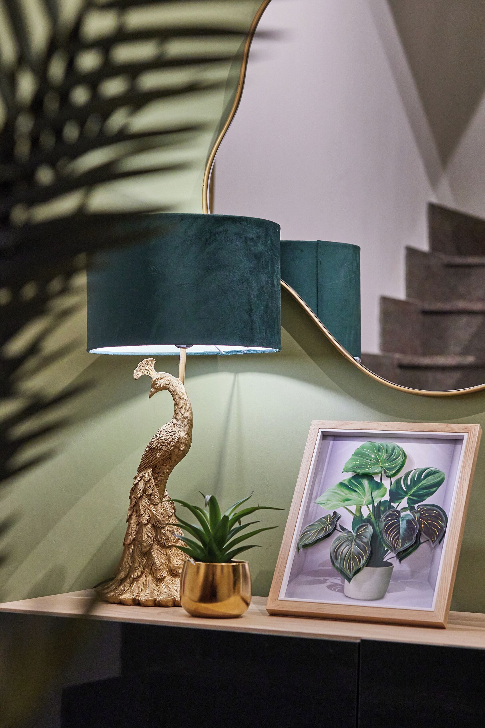 Une lampe ornée d’un motif de paon, une plante en pot encadrée et un miroir posé sur une étagère.