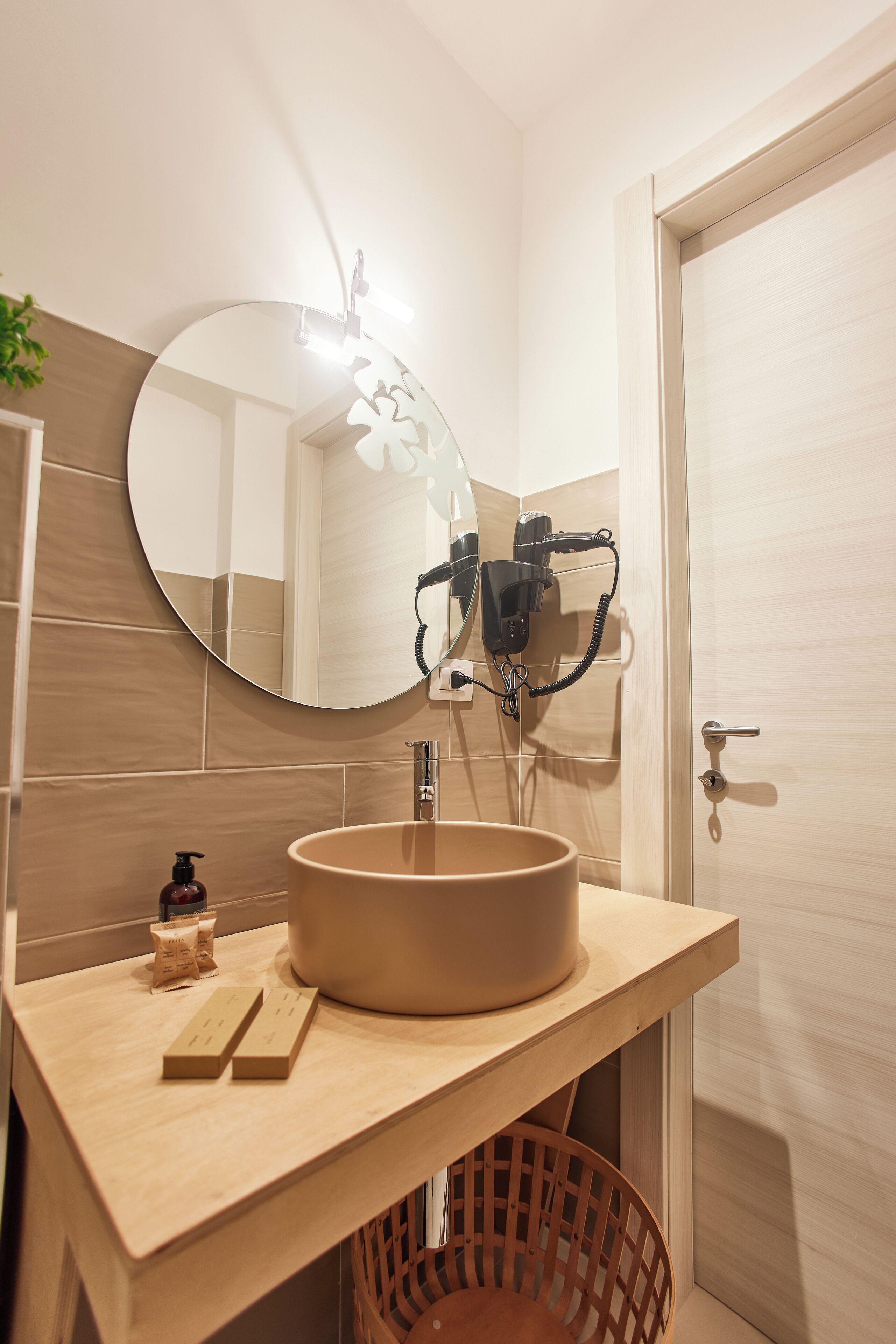 A bathroom with a round mirror, beige tiles, a sink, and a hairdryer on the wall.
