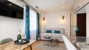 A modern living room with a sofa, two small side tables, a wooden dining table with a bottle and glass, and a kitchen area with a sink. - Furnished Studio La Jungle Suite, Mandara Beach (Antibes)