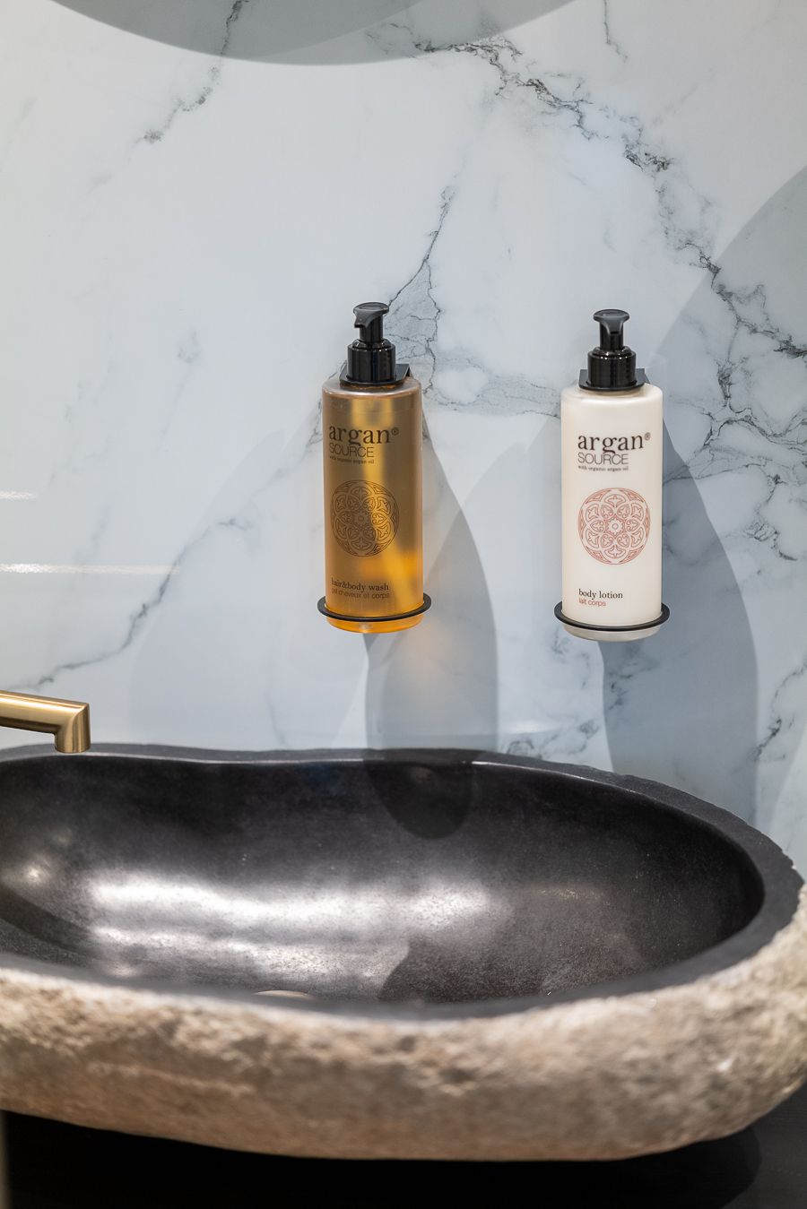 A bathroom with a marble wall, a black basin, and two Argan Source products.