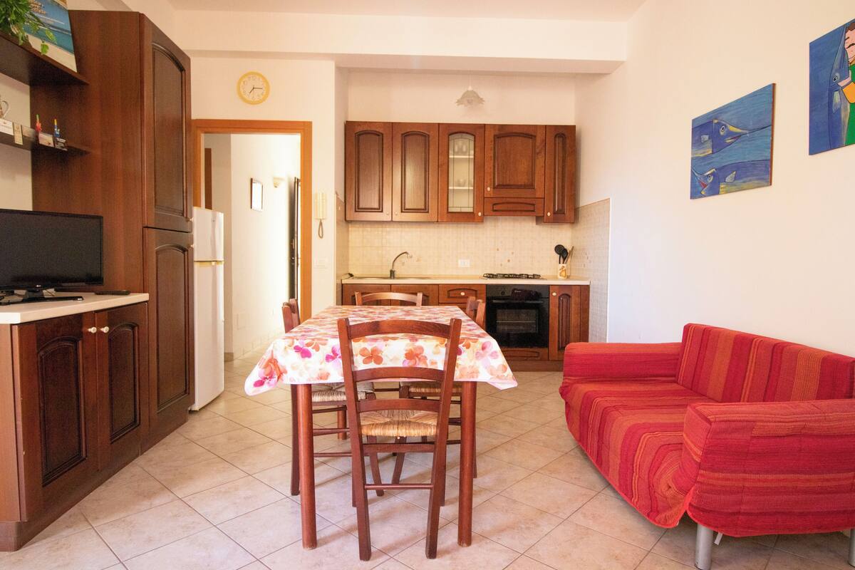 A compact kitchen with wooden cabinets, a dining table with a floral tablecloth, and a red sofa.