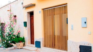 Un edificio con una puerta de madera grande, una ventana pequeña y una planta en maceta con flores rosadas.