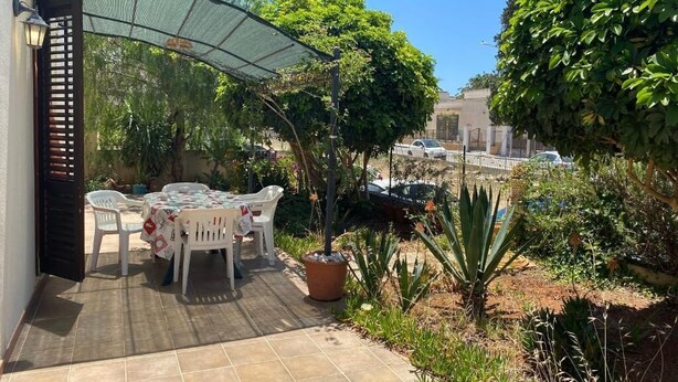 Un patio al aire libre con mesa y sillas, rodeado de follaje y un coche al fondo.