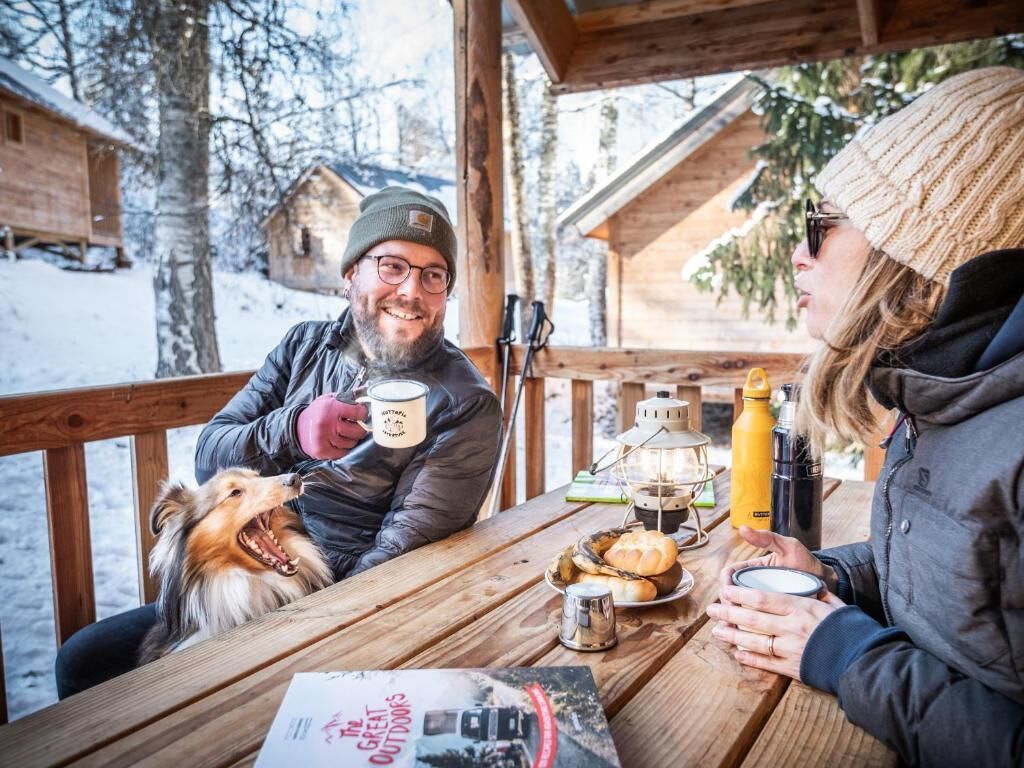 Una terraza de madera con una mesa preparada para dos personas y un perro, rodeada de árboles cubiertos de nieve.
