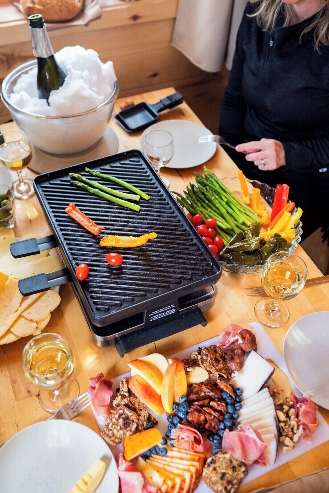 Una mesa con una parrilla, varias verduras, frutas, quesos, carnes y una botella de champán.