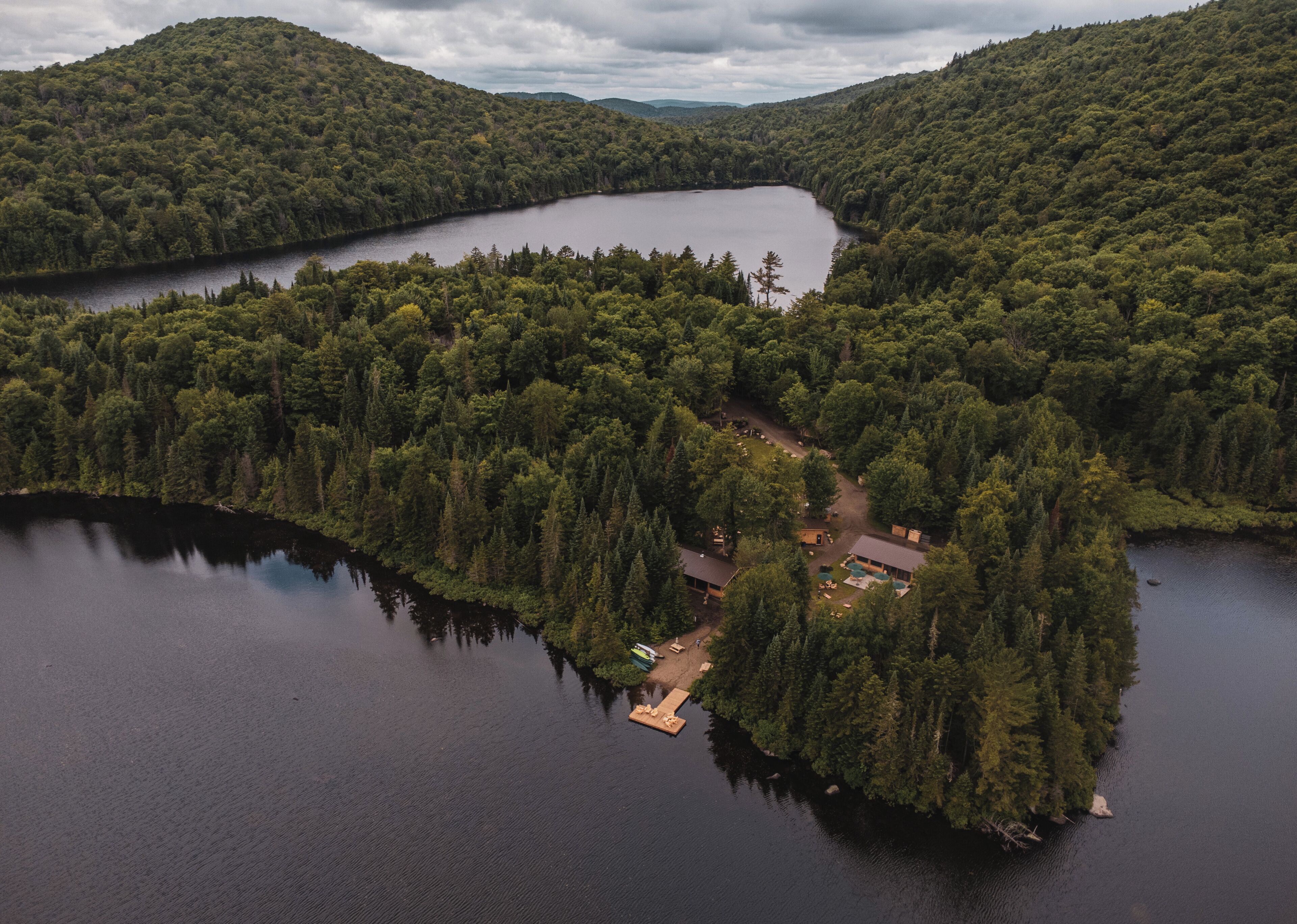 Vue aérienne d’une propriété au bord du lac, entourée d’une forêt dense, avec une jetée et un petit bâtiment doté d’une piscine.