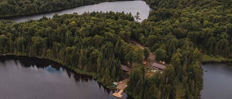 Vue aérienne d’une propriété au bord du lac, entourée d’une forêt dense, avec une jetée et un petit bâtiment doté d’une piscine.