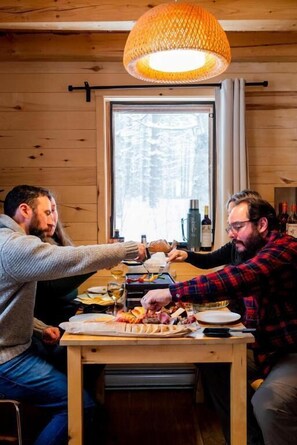 A wooden interior with a dining table, two people eating, and a window with a view of trees.