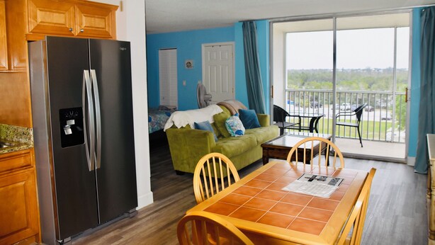 A compact living space with a kitchen area, a dining table with chairs, a green sofa, and a balcony with a view. - Easy Breezy Too! (Bonita Springs)