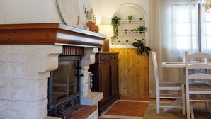 A cozy room with a fireplace, wooden beams, a dining area with chairs, and a kitchen counter with plants. - Casa Marmeleiro (Santiago do Cacém)