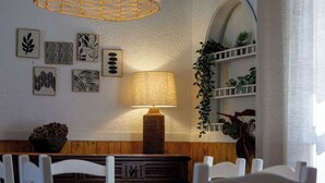 A dining area with a round table, white chairs, a woven pendant light, a wall-mounted lamp, and framed botanical prints. - Casa Marmeleiro (Santiago do Cacém)