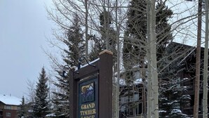 Un panneau enneigé indiquant l’emplacement du Grand Timber Lodge, avec une base en pierre, entouré d’arbres et de bâtiments.