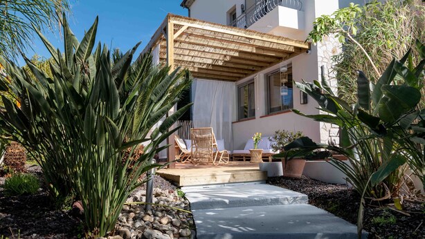 A patio area with a wooden pergola, outdoor seating, and tropical plants.