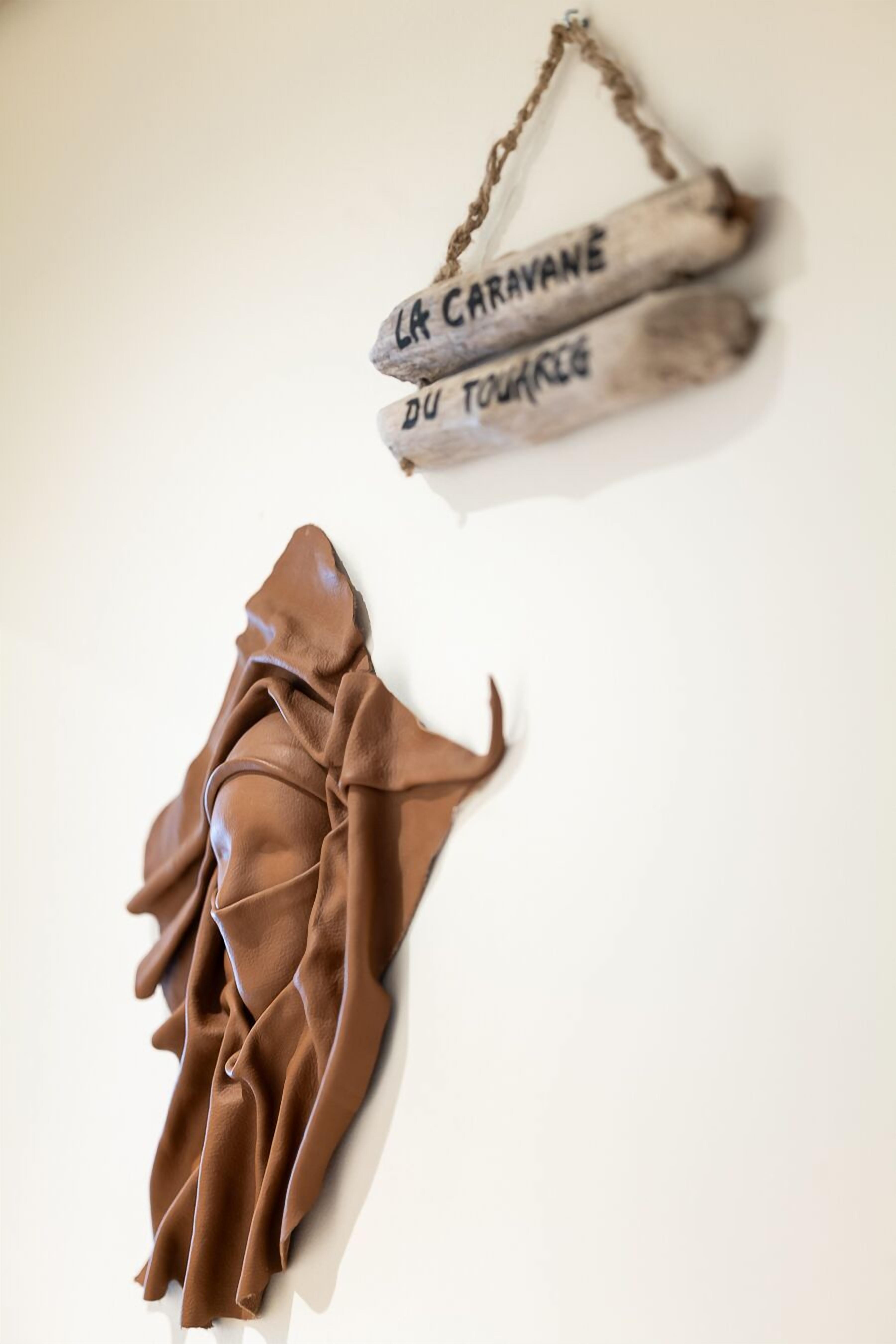 Una pared blanca con dos elementos colgantes: dos tablas de madera con la leyenda "LA CARAVANE DU TOURNOIS" y un trozo de tela marrón cubriendo una superficie.