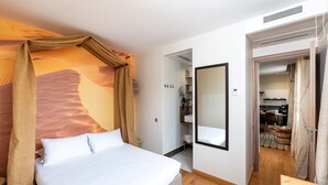 A hotel room with a bed, a mirror, and a view of a kitchen and dining area through an open door.