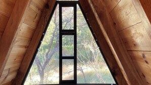 Un dormitorio rústico en el ático con un gran ventanal triangular, una cama con almohadas y vistas a árboles.