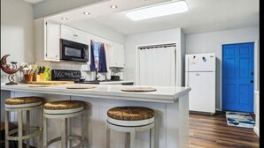 Una cocina con armarios blancos, microondas, refrigerador y una puerta azul. Hay tres taburetes con asientos de mimbre y una encimera blanca.
