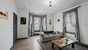 A living room with a sofa, a coffee table, and a rug. There are two windows with curtains, a door, and a chandelier.