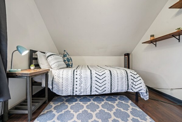 A bedroom with a bed, a desk with a lamp, a small plant, a patterned rug, and a wooden shelf with a plant. - Charming 6-bedroom house - close to all in Providence (Providence)