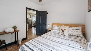 A bedroom with a bed, a wooden headboard, a ceiling fan, a nightstand, a small table with a plant, and a door leading to a kitchen.