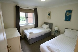A hotel room with two beds, a window with curtains, a bedside table with a lamp, and a picture on the wall.