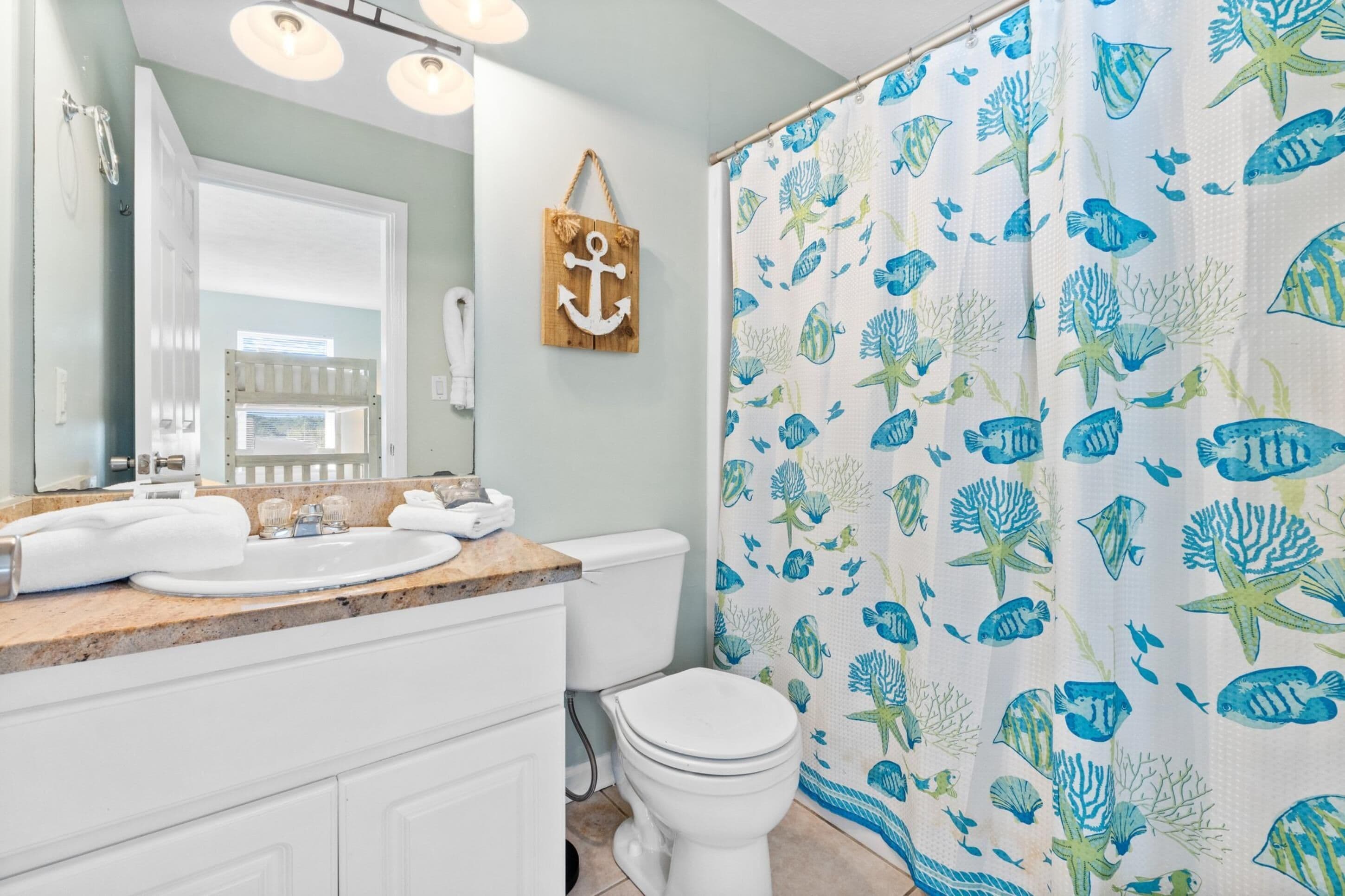 A bathroom with a white sink, mirror, and toilet. There is a shower curtain with a fish and coral pattern.