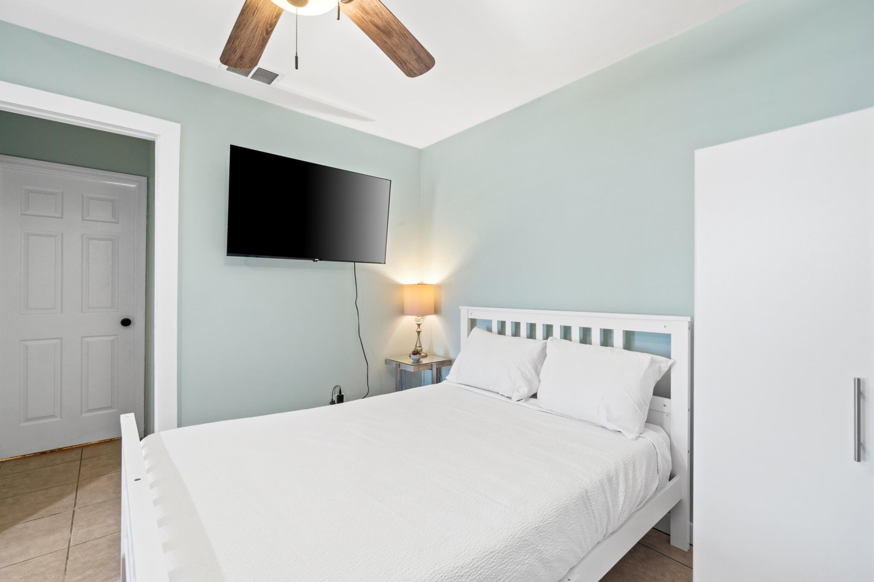 Een slaapkamer met een bed, een tv aan de muur, een plafondventilator en een nachtkastje met een lamp.