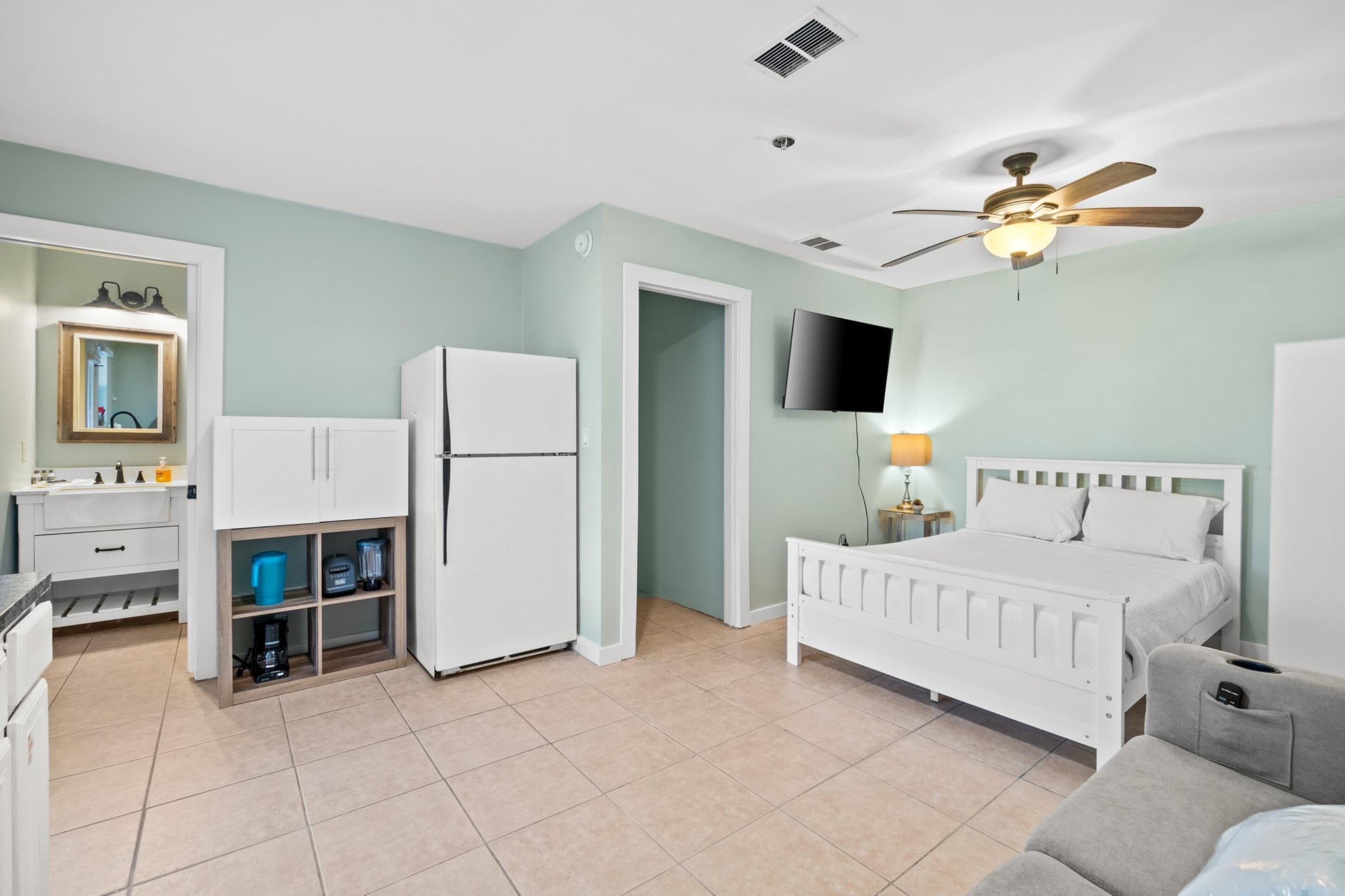 A bedroom with a bed, a ceiling fan, a TV, and a small kitchenette with a refrigerator and a coffee maker.
