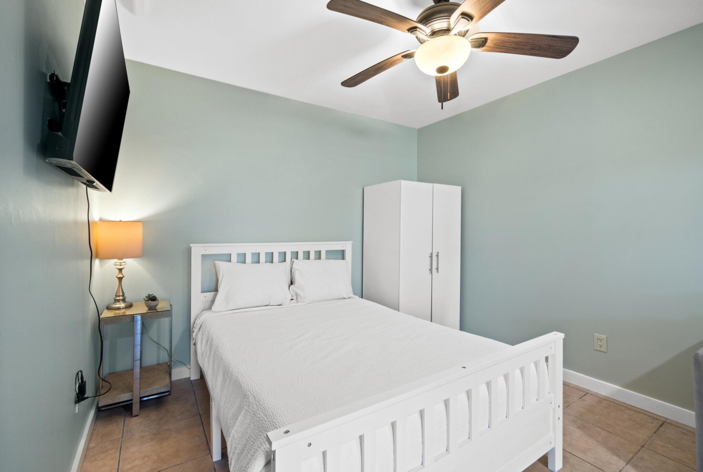 A bedroom with a bed, a wardrobe, a ceiling fan, a TV mounted on the wall, and a bedside table with a lamp.