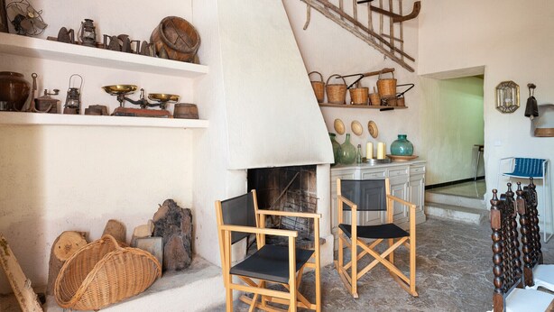 A rustic interior with wooden shelves displaying vintage kitchenware, a fireplace, and a dining area with wooden chairs.